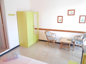 Um quarto com um guarda-roupa verde, duas cadeiras de vime, uma mesa pequena e uma cama.