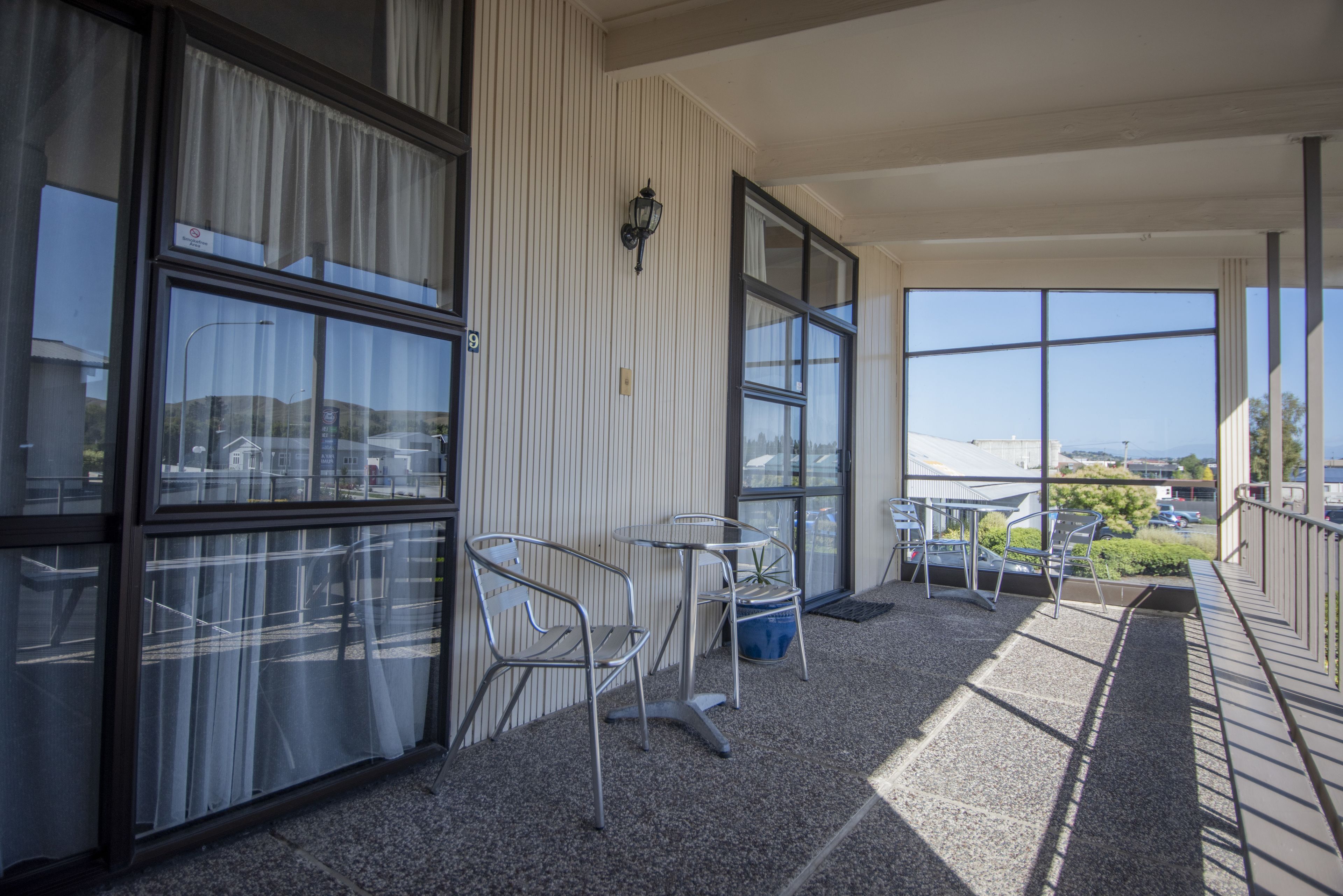 A covered balcony with metal chairs and a table, large windows, and a view of the surrounding area.