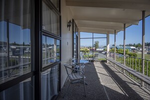 Uma área externa coberta com cadeiras e mesas de metal, grandes janelas e vista para um estacionamento e edifícios.