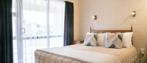 Una camera d'albergo con un letto grande, una finestra con tende leggere, un muro a mattoni e un comodino con una lampada.
