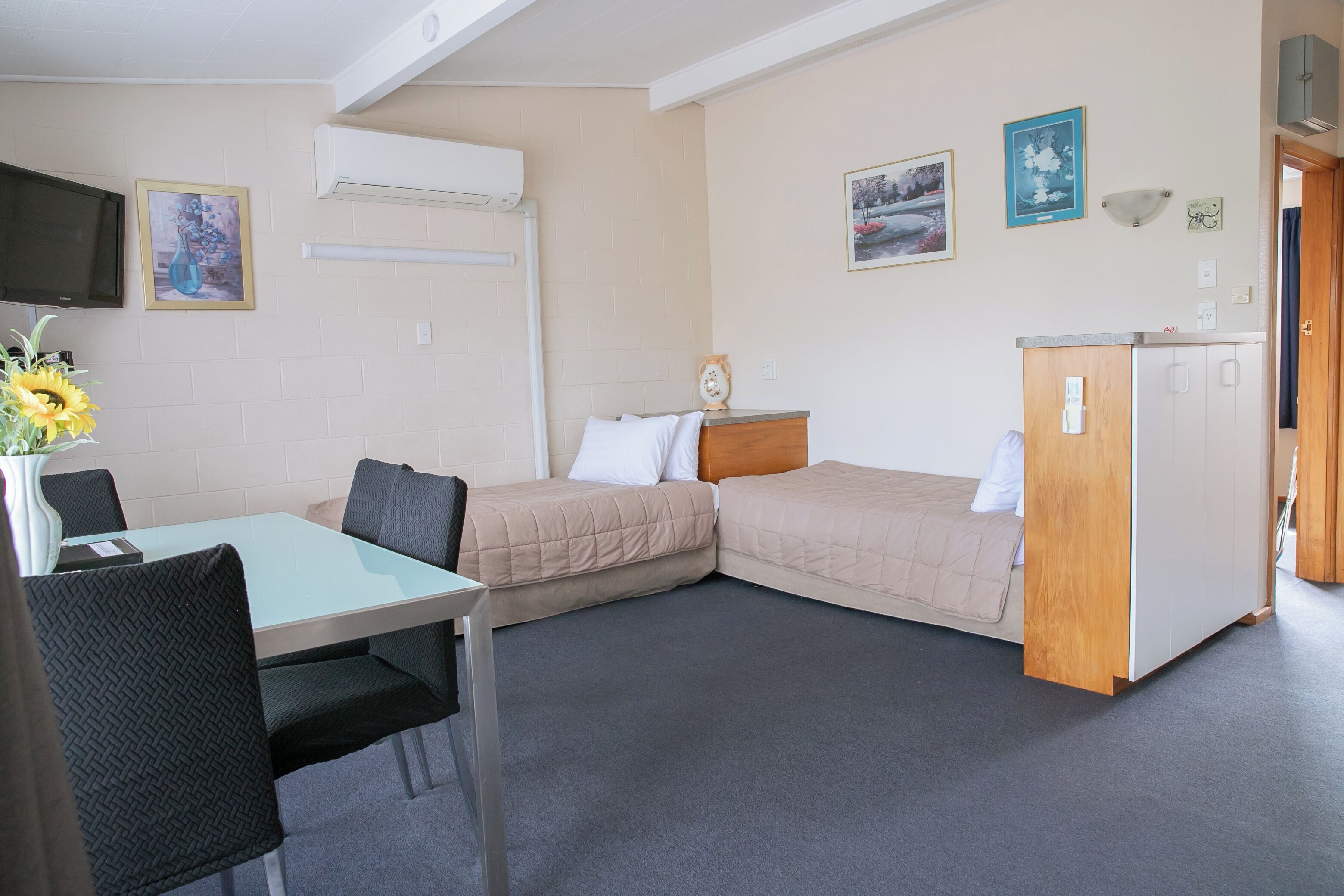 A room with a bed, a desk, a chair, a small table with a vase of flowers, and a TV mounted on the wall.