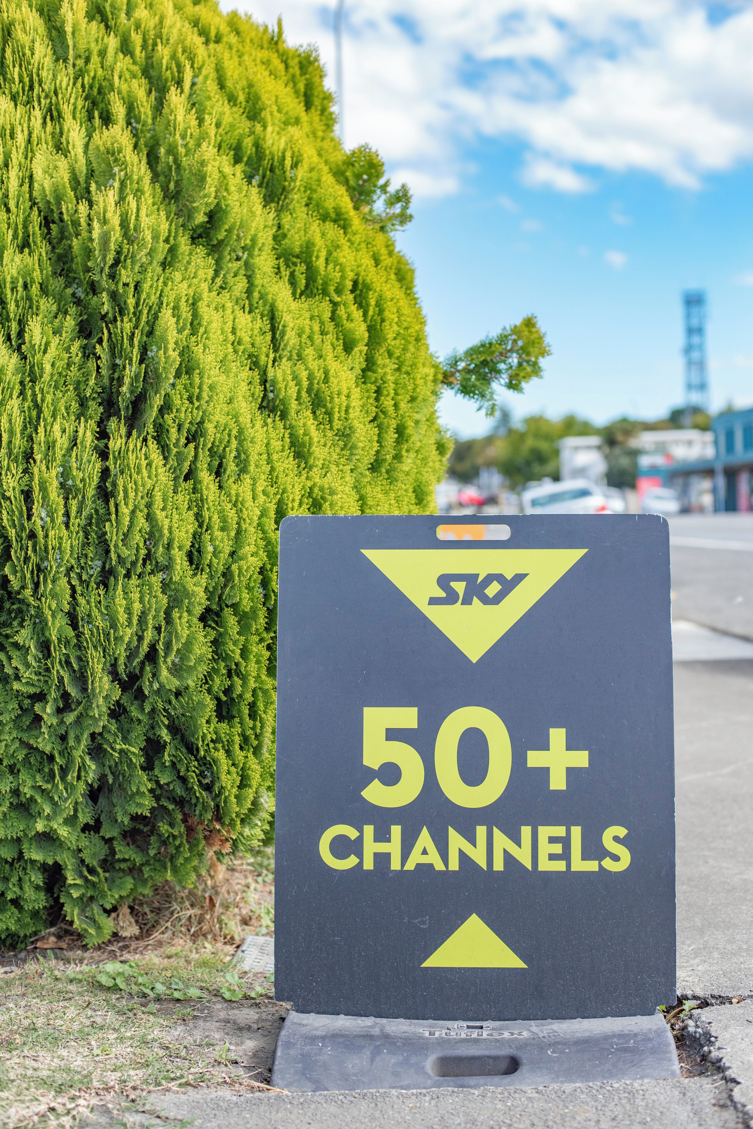 A sign with the SKY logo and "50+ CHANNELS" text, placed on a concrete base beside a hedge.