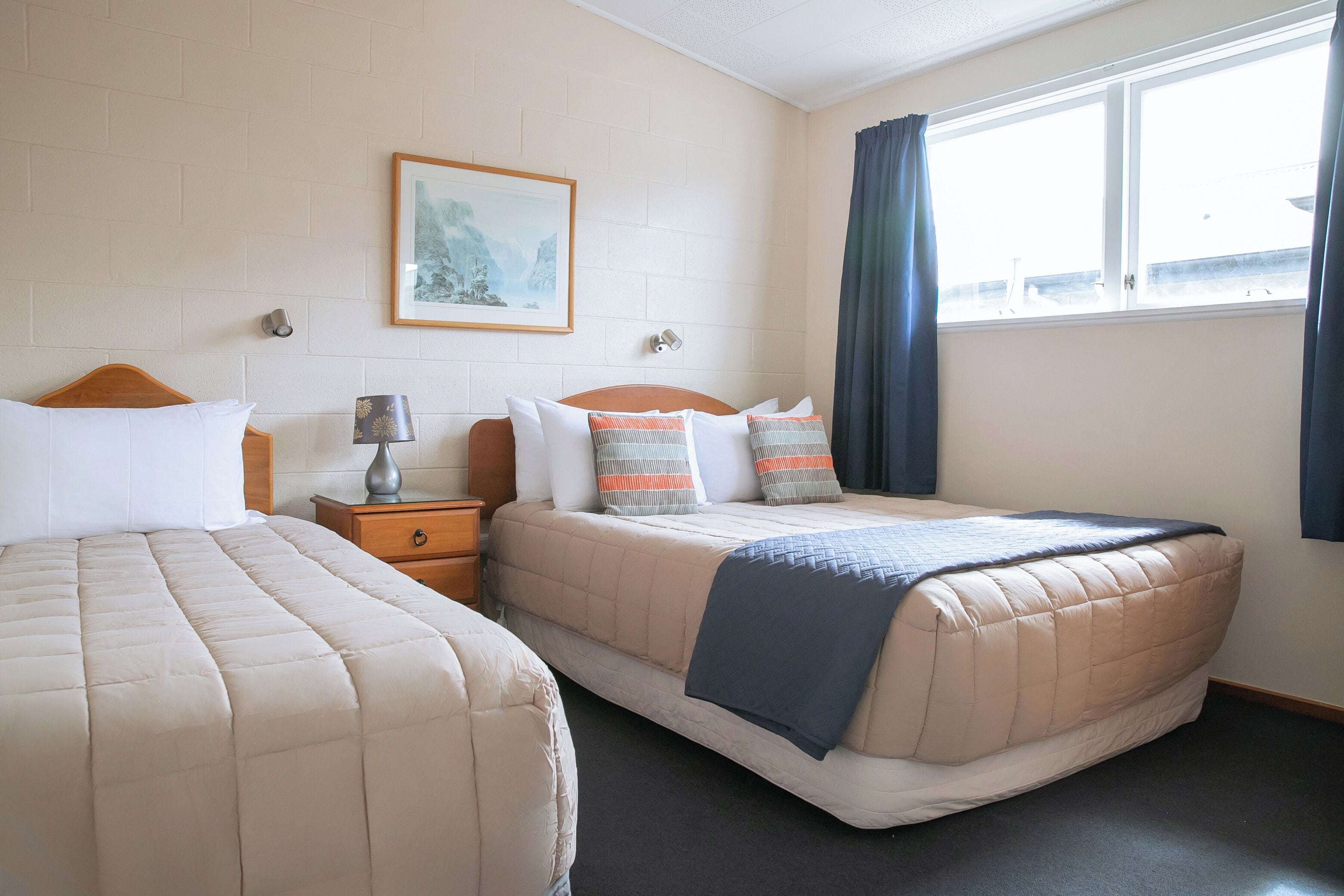 Une chambre d’hôtel avec deux lits, une table de chevet, une lampe, un tableau au mur et une fenêtre avec des rideaux.