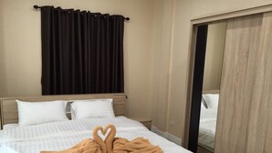 Une chambre d’hôtel avec un lit, deux oreillers, une serviette marron disposée en forme de cœur, un rideau foncé et un miroir sur le mur.