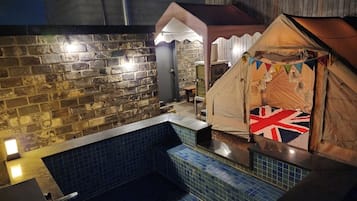 Un espace extérieur entouré de murs en pierre, avec une piscine carrelée, une tente ornée d’un drapeau Union Jack et une structure en bois.