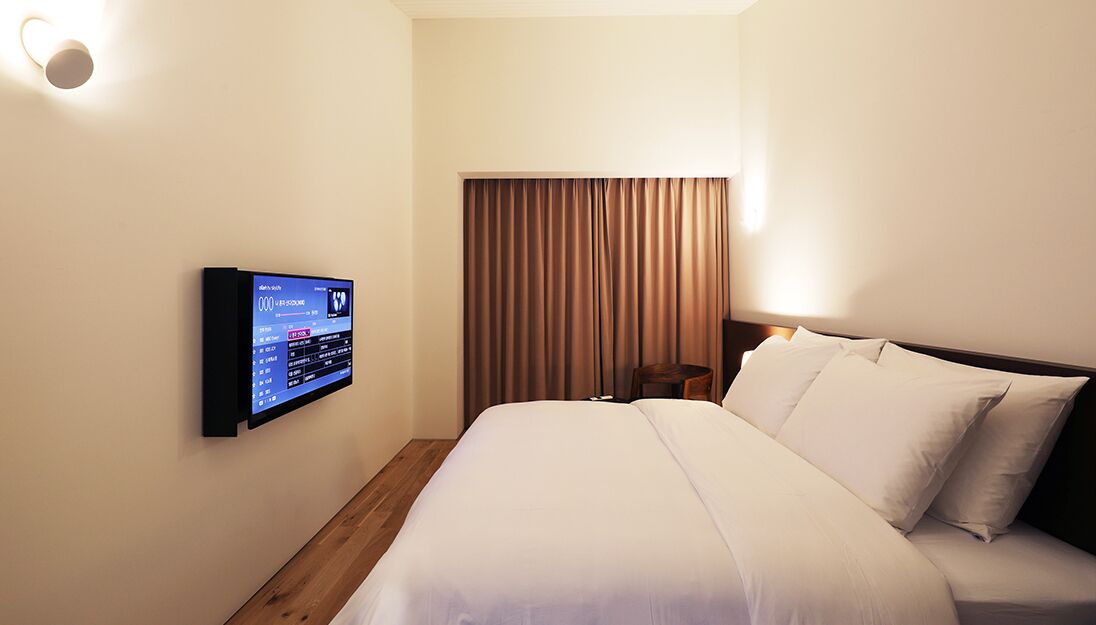 A hotel room with a bed, a wall-mounted control panel, and a window with curtains.
