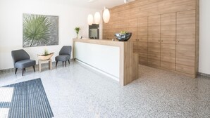 A modern reception area with a wooden counter, two grey chairs, a large abstract painting, and a potted plant. - Ambient Hotel Tassilo (Dingolfing)