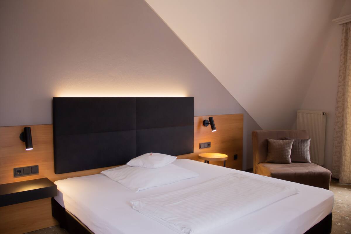 A modern hotel room with a large bed, a nightstand, a wall-mounted lamp, and a small round table.