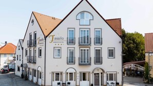 A two-story building with a white facade and a brown roof, labeled as "Jassilo Ambienthotel." It features multiple windows, balconies, and a small outdoor seating area. - Ambient Hotel Tassilo (Dingolfing)