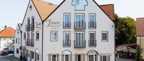 A two-story building with a white facade and a brown roof, labeled as "Jassilo Ambienthotel." It features multiple windows, balconies, and a small outdoor seating area.
