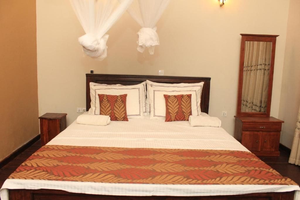 A double bed with a patterned bedspread, two pillows with a leaf design, a nightstand, a wooden cabinet, and a mirror with a curtain.