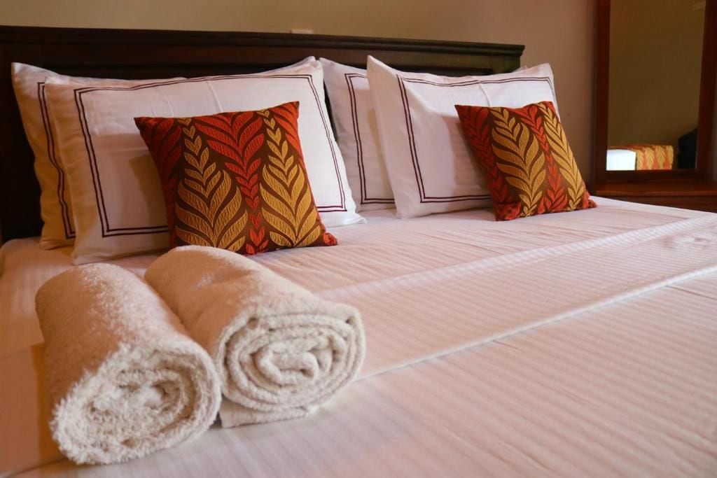 Quarto de hotel com cama bem arrumada, duas almofadas com padrão de folha e dois rolinhos de toalha.