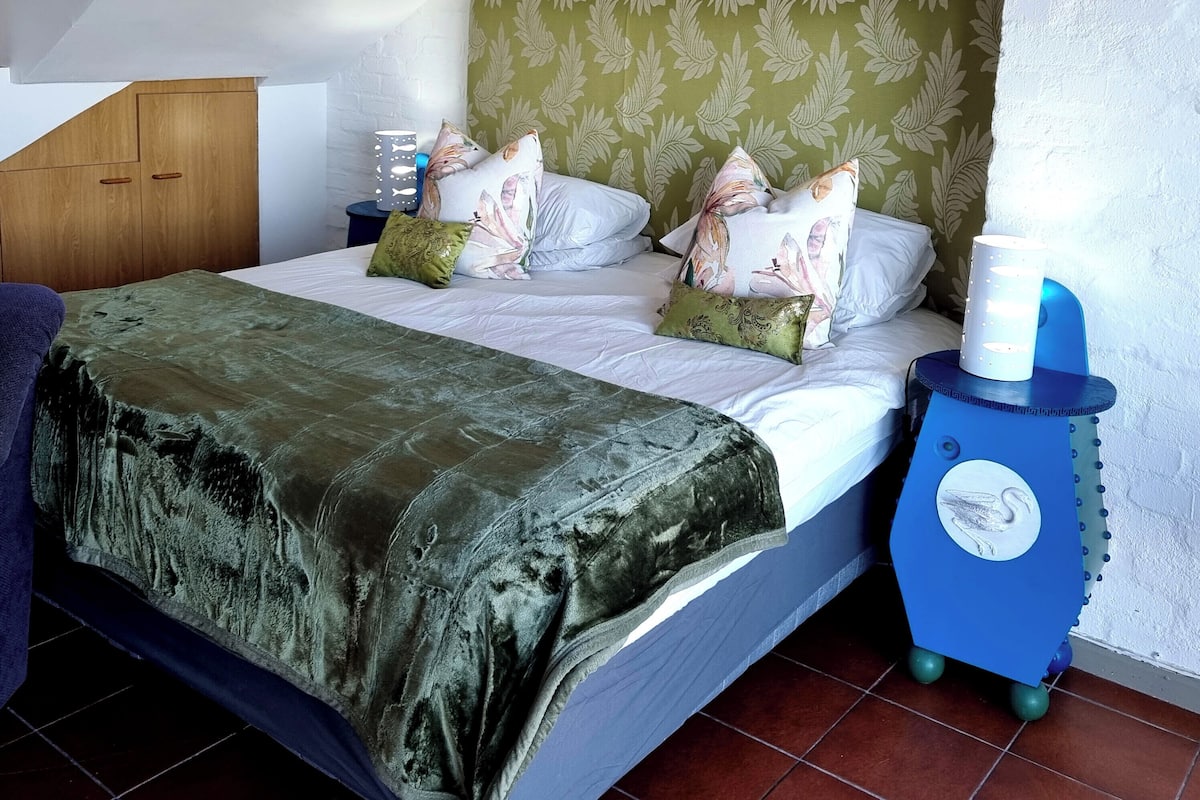 A bedroom with a bed, pillows, a nightstand, a lamp, and a blue bedside table with a clock.