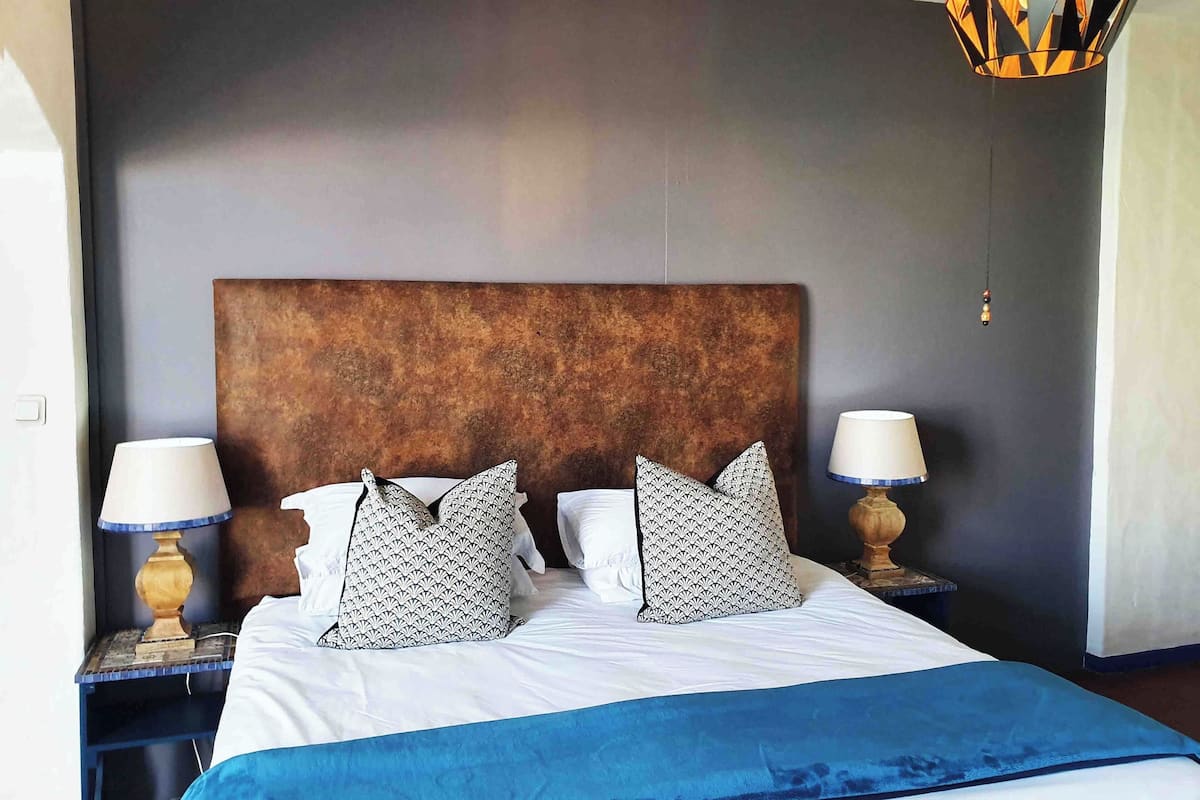 A neatly made bed with patterned pillows, a blue bedspread, and a textured brown headboard. There are two bedside lamps with white shades and a geometric-patterned hanging light fixture.