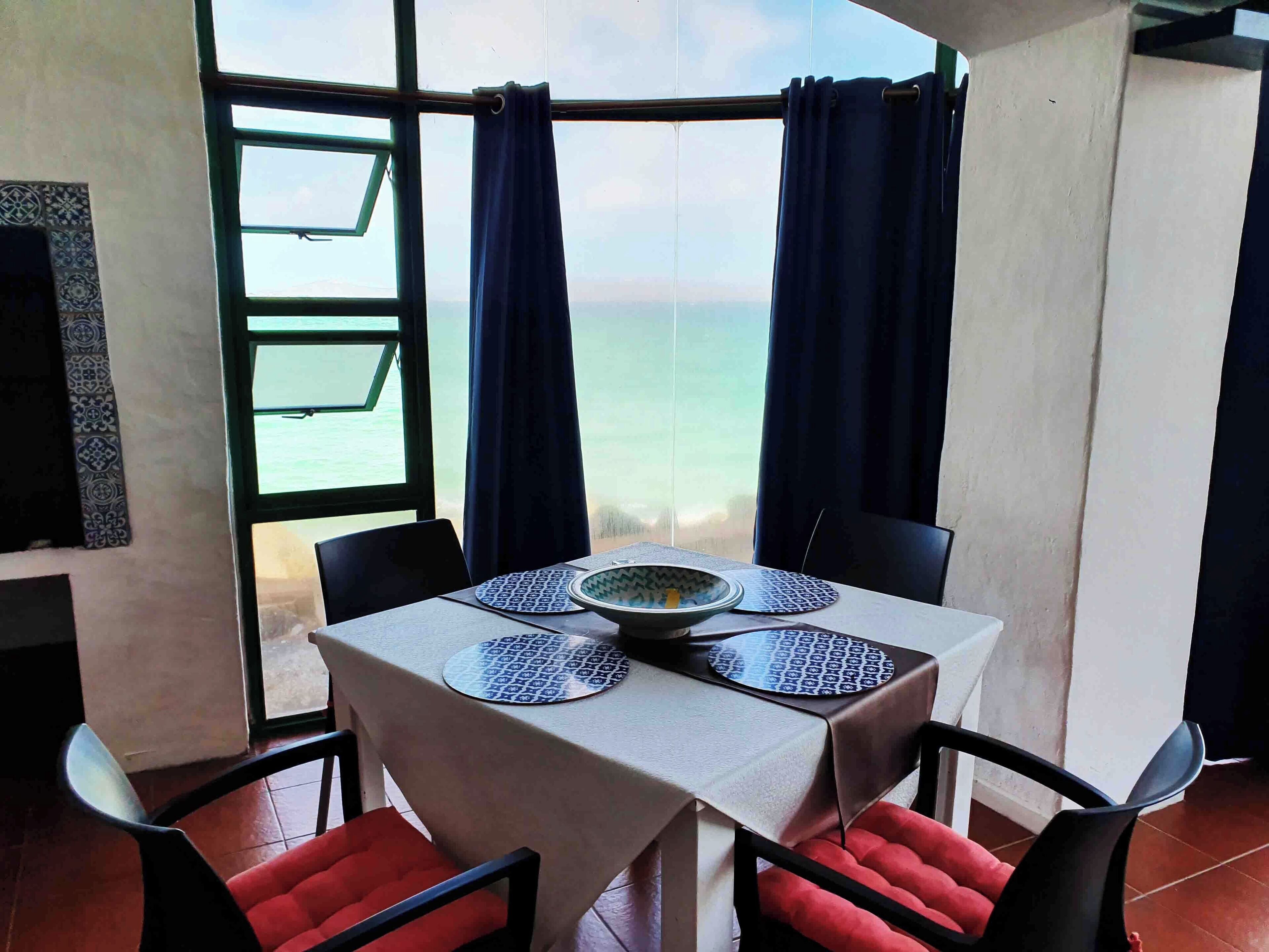 A dining area with a table set for two, near a large window overlooking the sea.