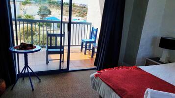 Habitación de hotel con balcón, una cama con manta roja, una mesita con una bebida y una silla.