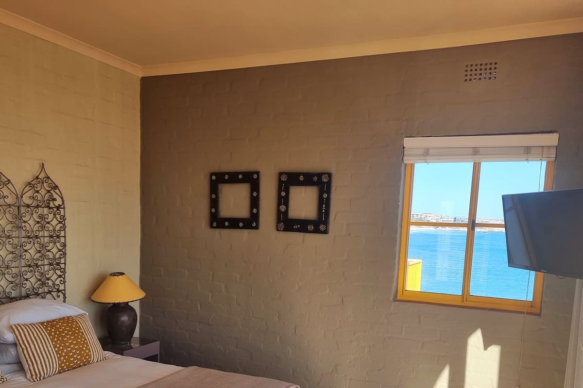 A bedroom with a bed, a lamp, two decorative frames, a TV, and a window with a view of the sea.