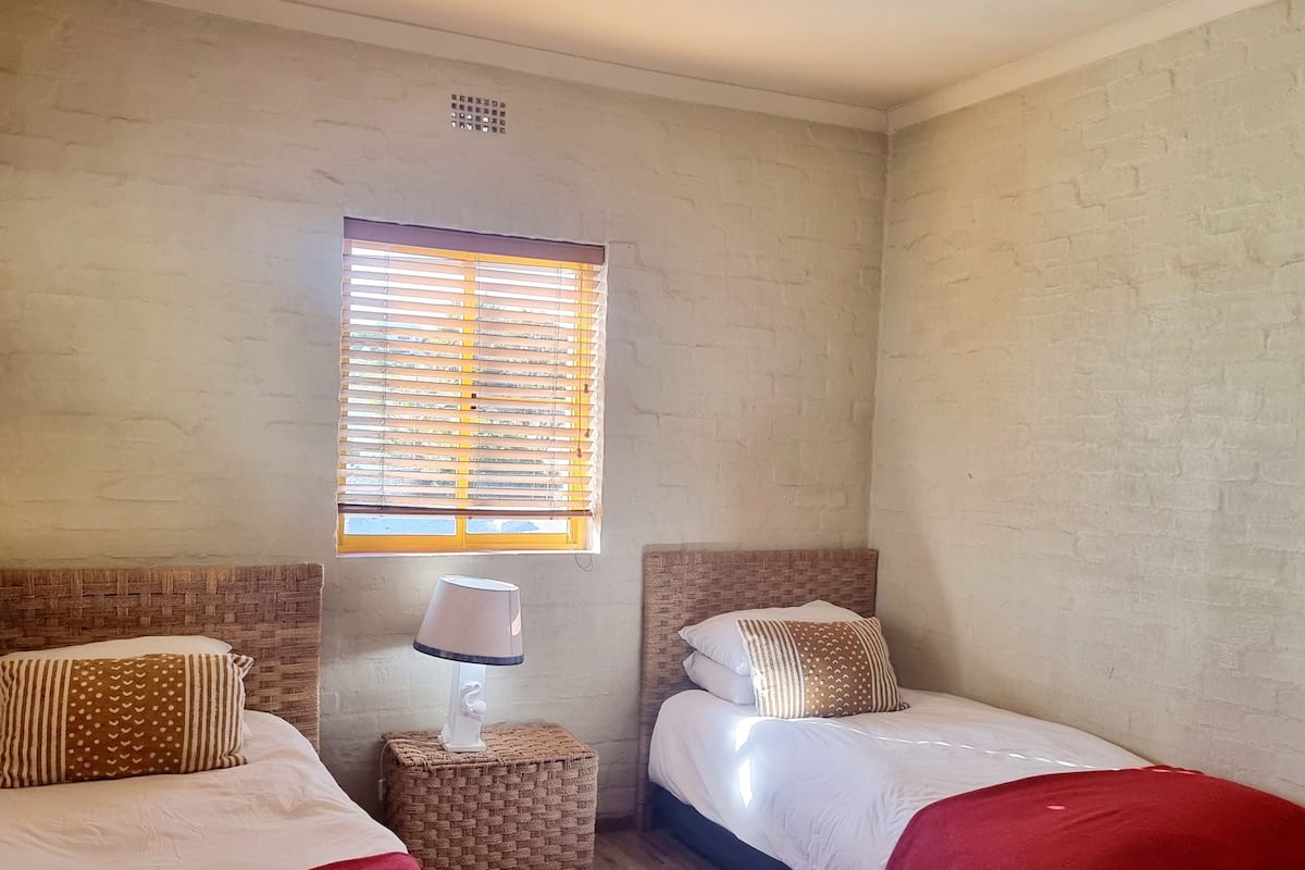A room with two beds, a small table with a lamp, a window with blinds, and a red rug.
