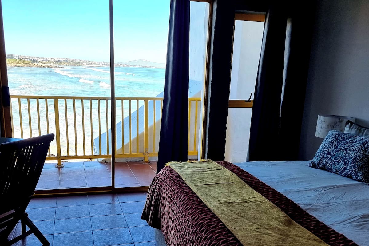 A bedroom with a large bed, a chair, and a balcony with a view of the sea.