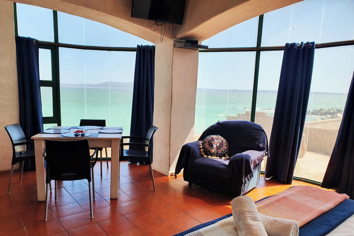 A room with a large window overlooking the sea, a bed with a rolled towel, a table with chairs, and a dark armchair with a patterned cushion.