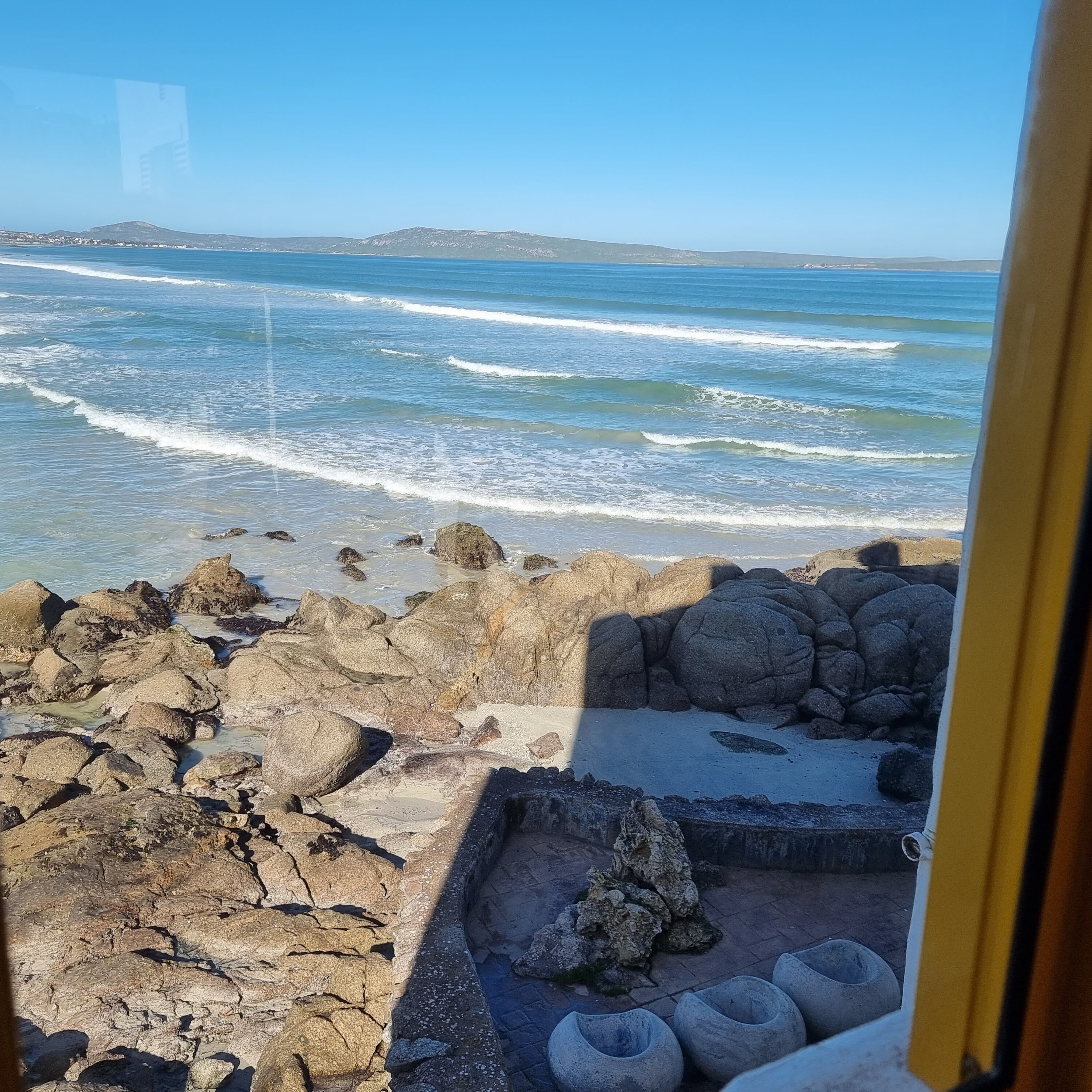Uma praia rochosa com ondas quebrando na costa, céu claro e montanhas ao longe.