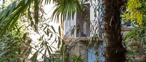 A view through dense foliage to a building with a balcony and a metal railing.