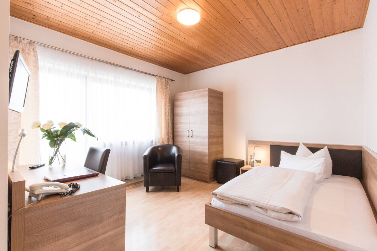 A hotel room with a wooden ceiling, a bed with white linens, a desk with a telephone, a chair, and a wardrobe.