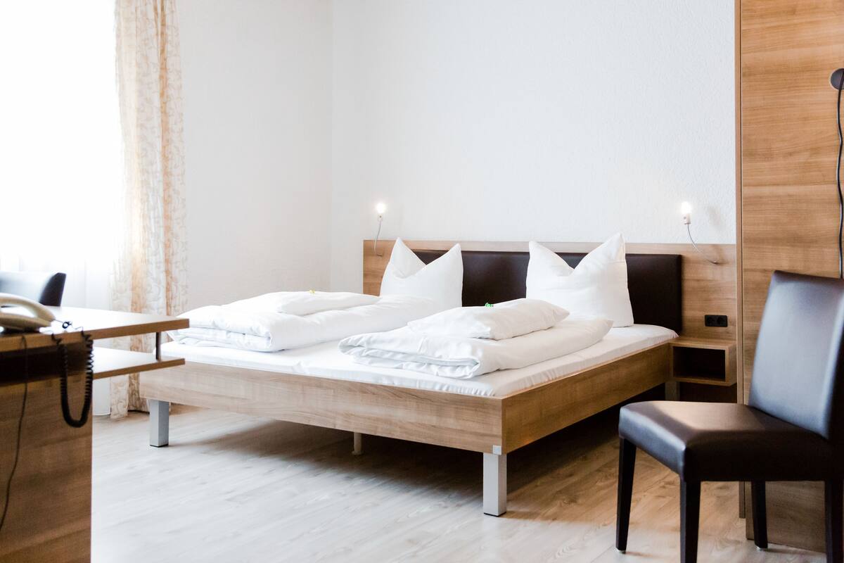 A neatly made bed with white linens, a wooden headboard, bedside tables with lamps, a desk with a telephone, and a chair.