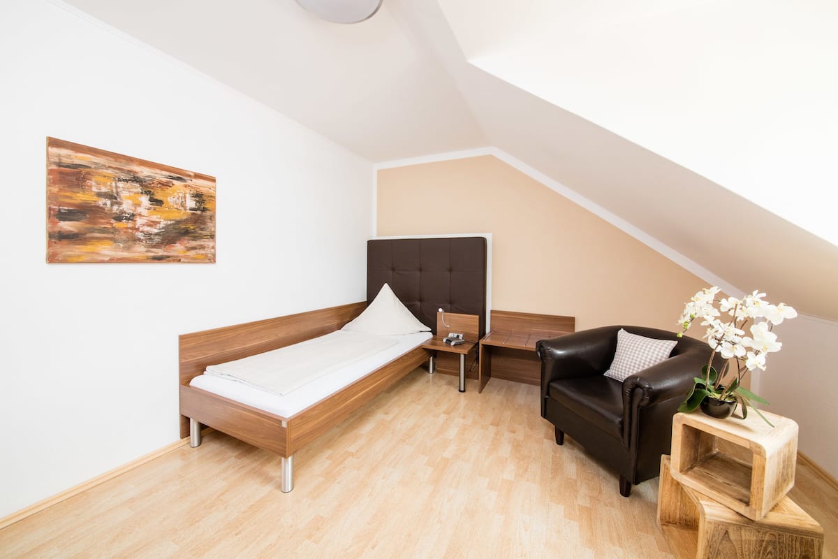 A bedroom with a wooden bed, a black leather chair, a small wooden nightstand, a white pillow, and a painting on the wall.