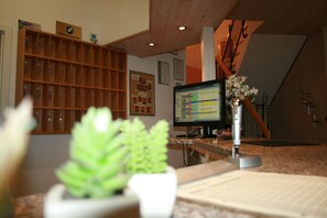 Un mostrador de recepción de hotel con un monitor de computadora, una planta y una estantería de madera con varios objetos.