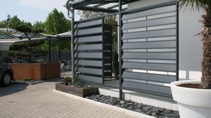 A modern building with a metal facade, a covered outdoor seating area, and a large planter with a tree. - Hotel Bavaria Garni (Dingolfing)