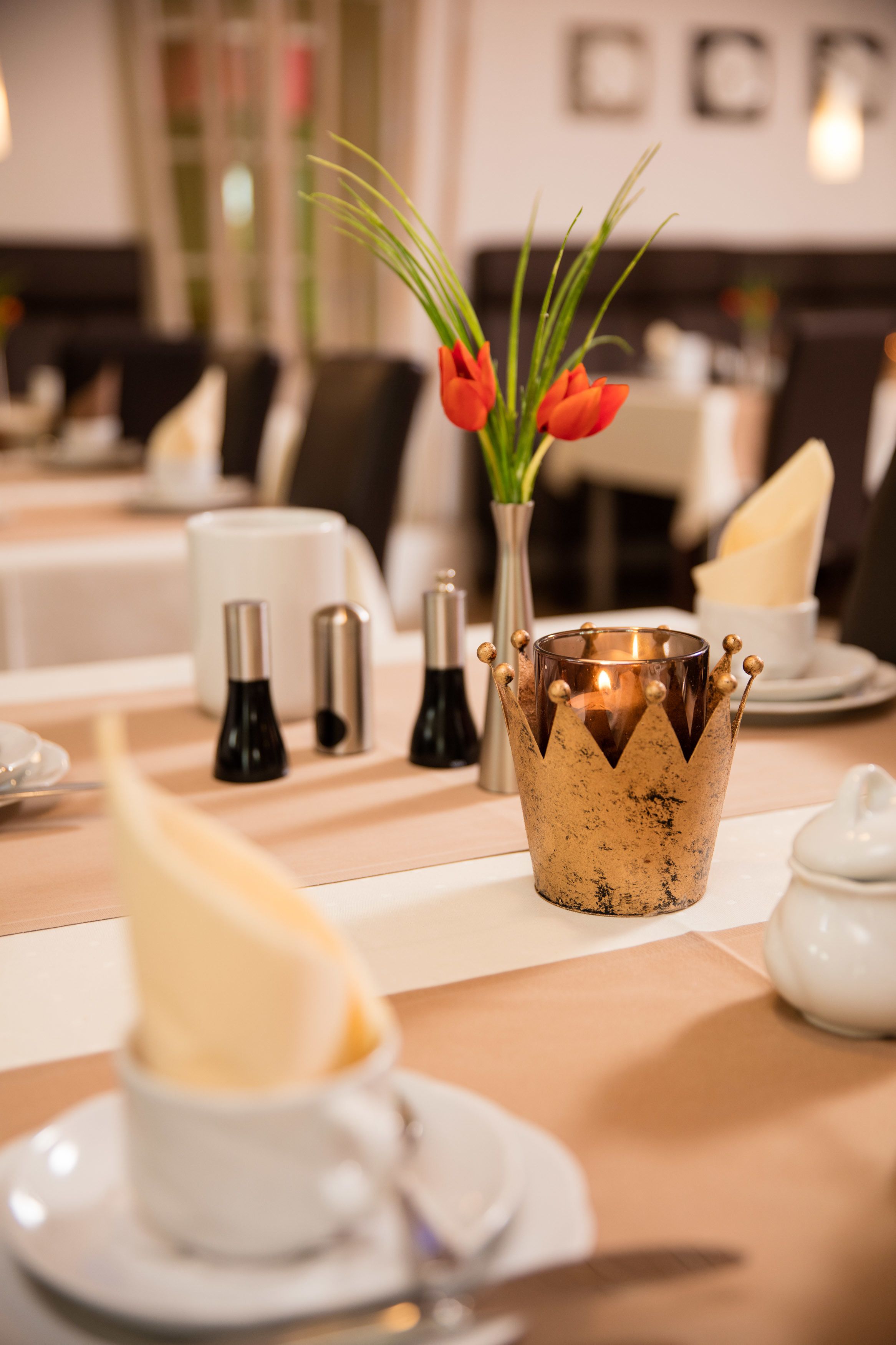Uma mesa de jantar com uma vela em castiçal decorativo, um vaso com tulipas vermelhas, saleiro e pimenteiro e uma chaleira branca.