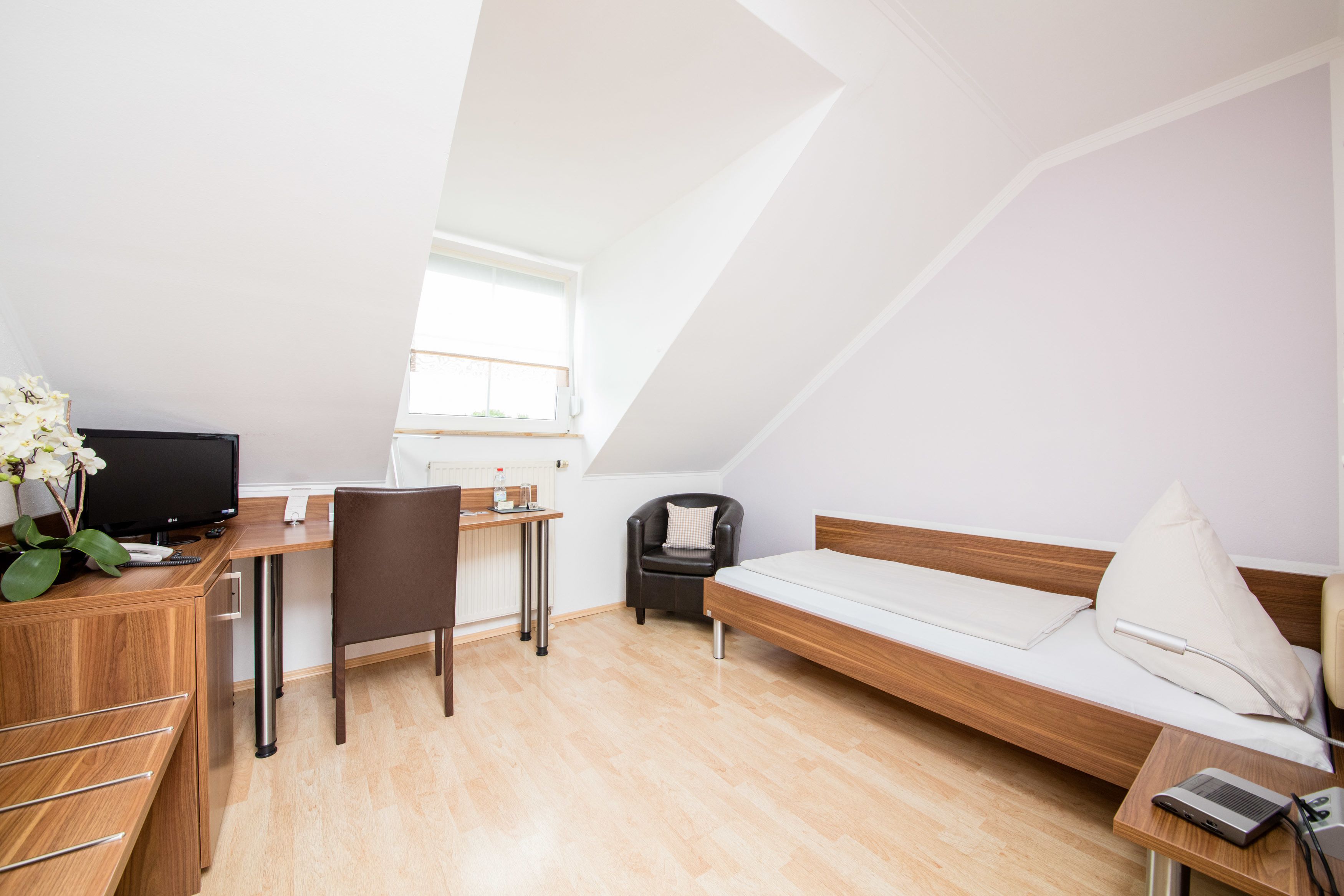 Una habitación moderna y bien iluminada con un escritorio de madera, una computadora, una silla, una cama con almohada y una mesita de noche con lámpara.