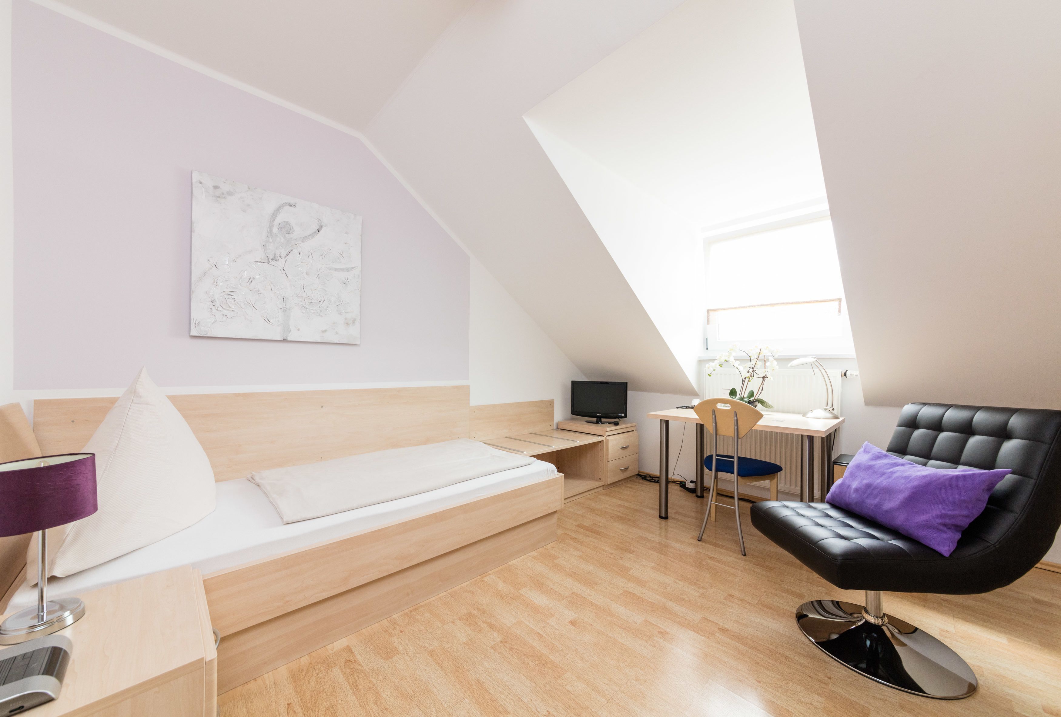 A modern loft bedroom with a sloped ceiling, a bed with a headboard, a desk with a chair, a black leather armchair, and a small table with a lamp.