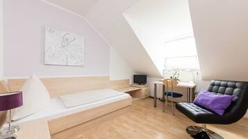 A modern loft bedroom with a sloped ceiling, a bed with a headboard, a desk with a chair, a black leather armchair, and a small table with a lamp.