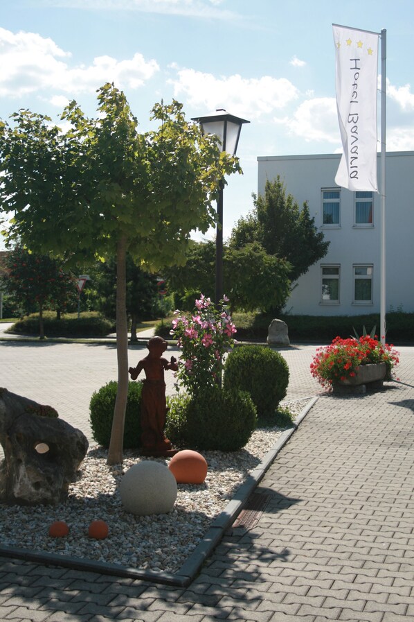 Un jardín ajardinado con una estatua, una bandera y un edificio al fondo.