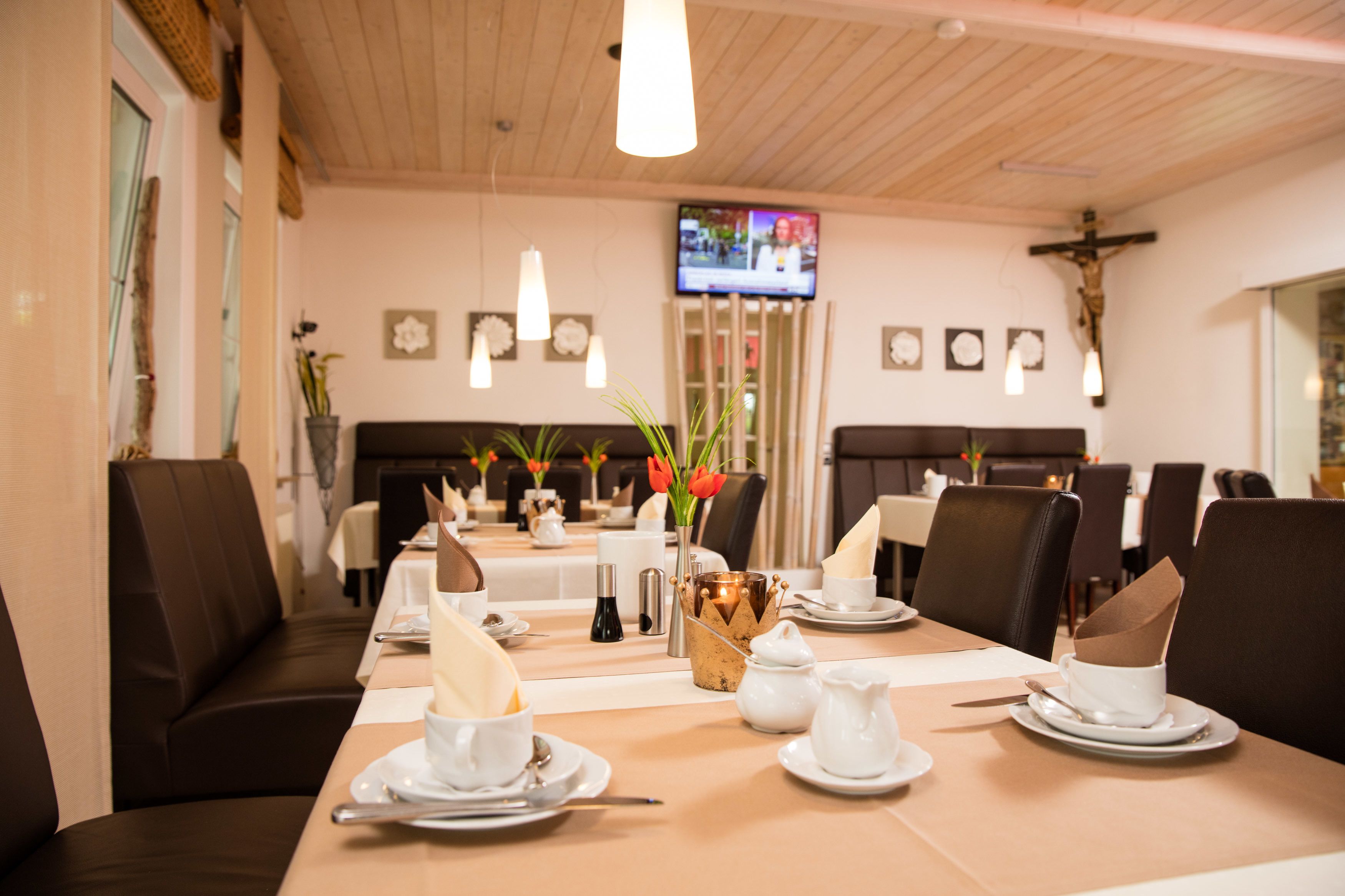 A dining area with tables set for a meal, a television mounted on the wall, and a cross on the wall.