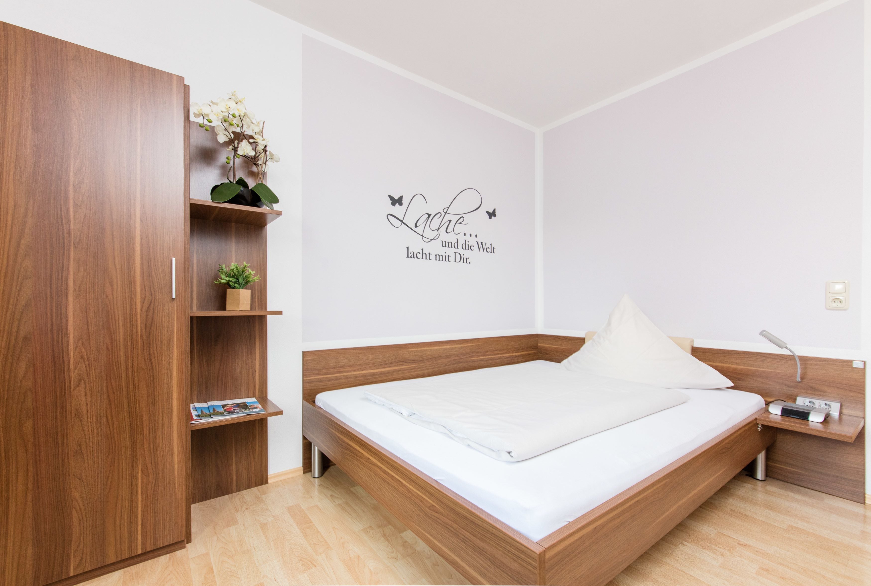 Un dormitorio moderno con un cabecero de madera, una mesita de noche, un estante con una planta y una pared con un letrero decorativo.