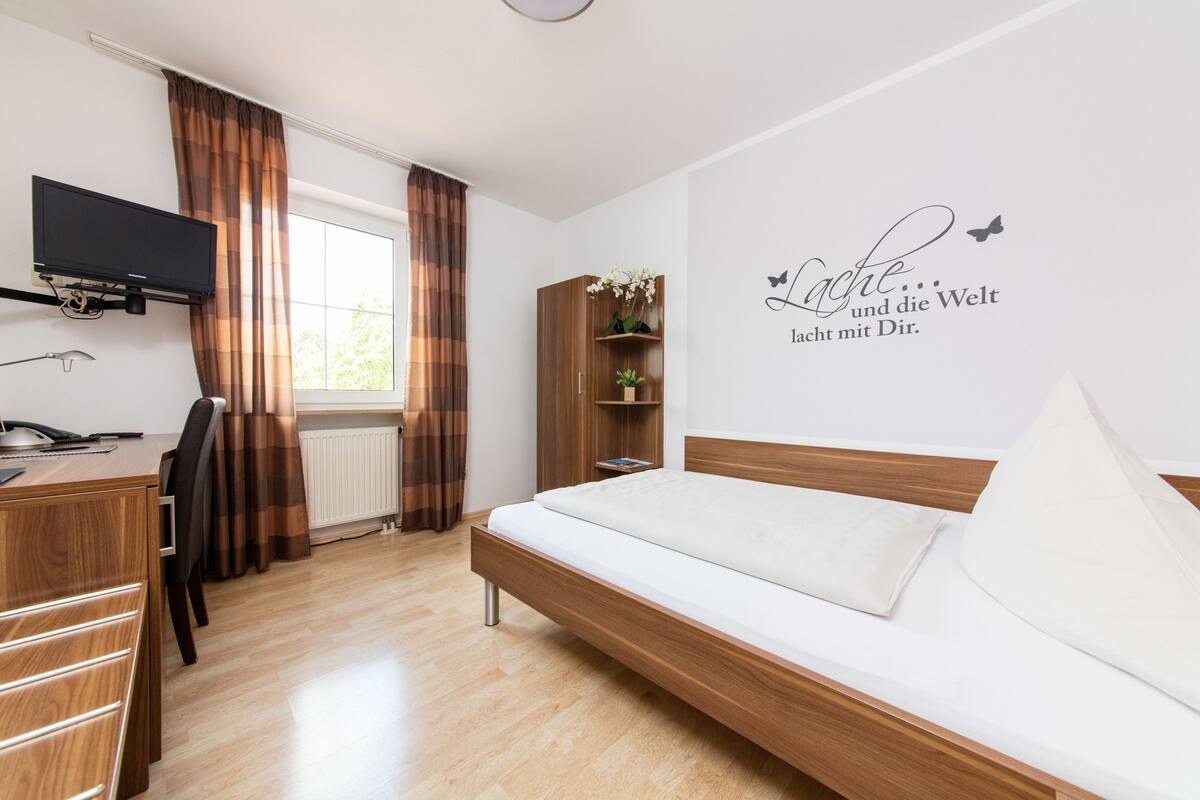 A hotel room with a wooden desk, a bed, a TV mounted on the wall, and a window with curtains.