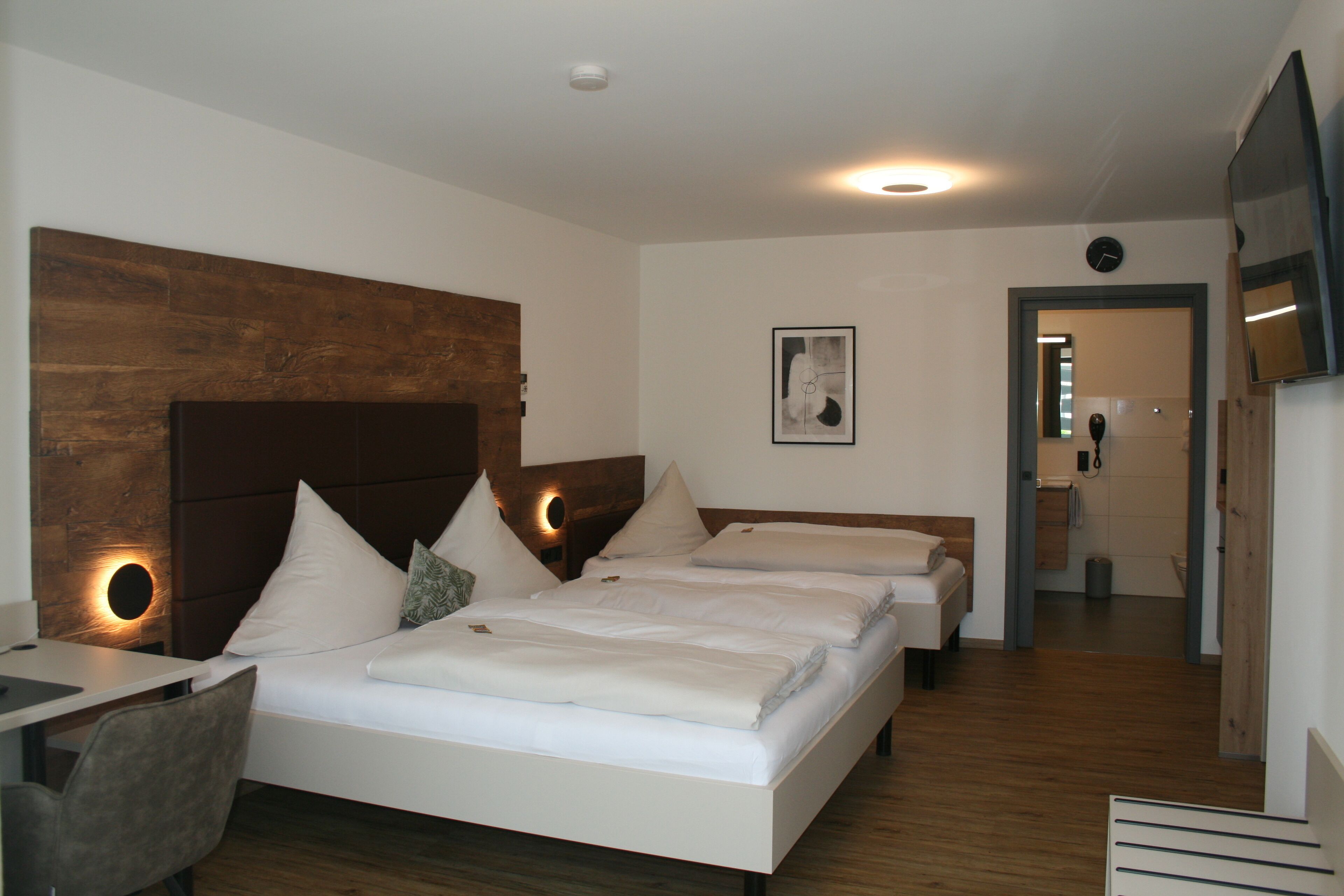 A hotel room with a large bed, a desk, and a chair. There is a wooden headboard, a wall clock, and a framed picture on the wall.