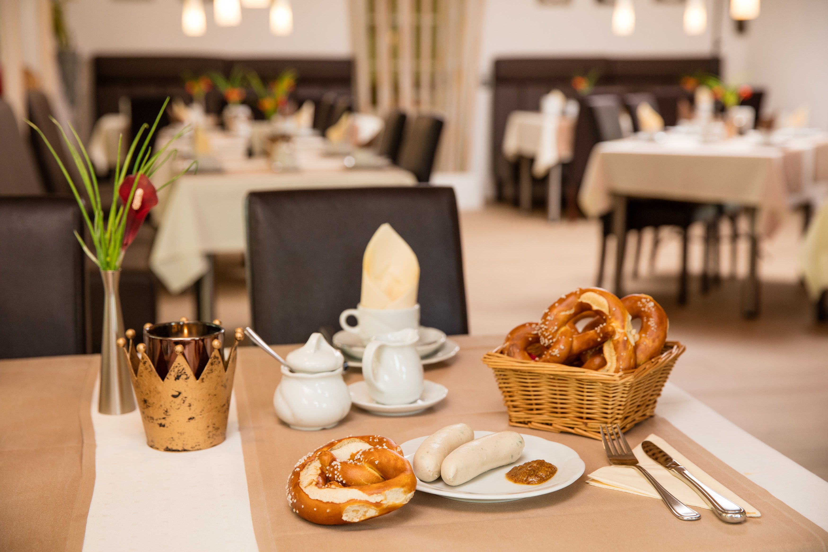 Ein Esstisch mit verschiedenen Speisen, darunter Brezeln, Würstchen und einem Korb mit Brot. Außerdem befinden sich kleine weiße Tassen und eine Metalltasse mit einem Wappentier darauf.