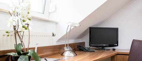 Un escritorio de madera con un monitor de computadora, un teléfono y una lámpara de escritorio. Hay una planta en maceta con flores blancas sobre el escritorio.