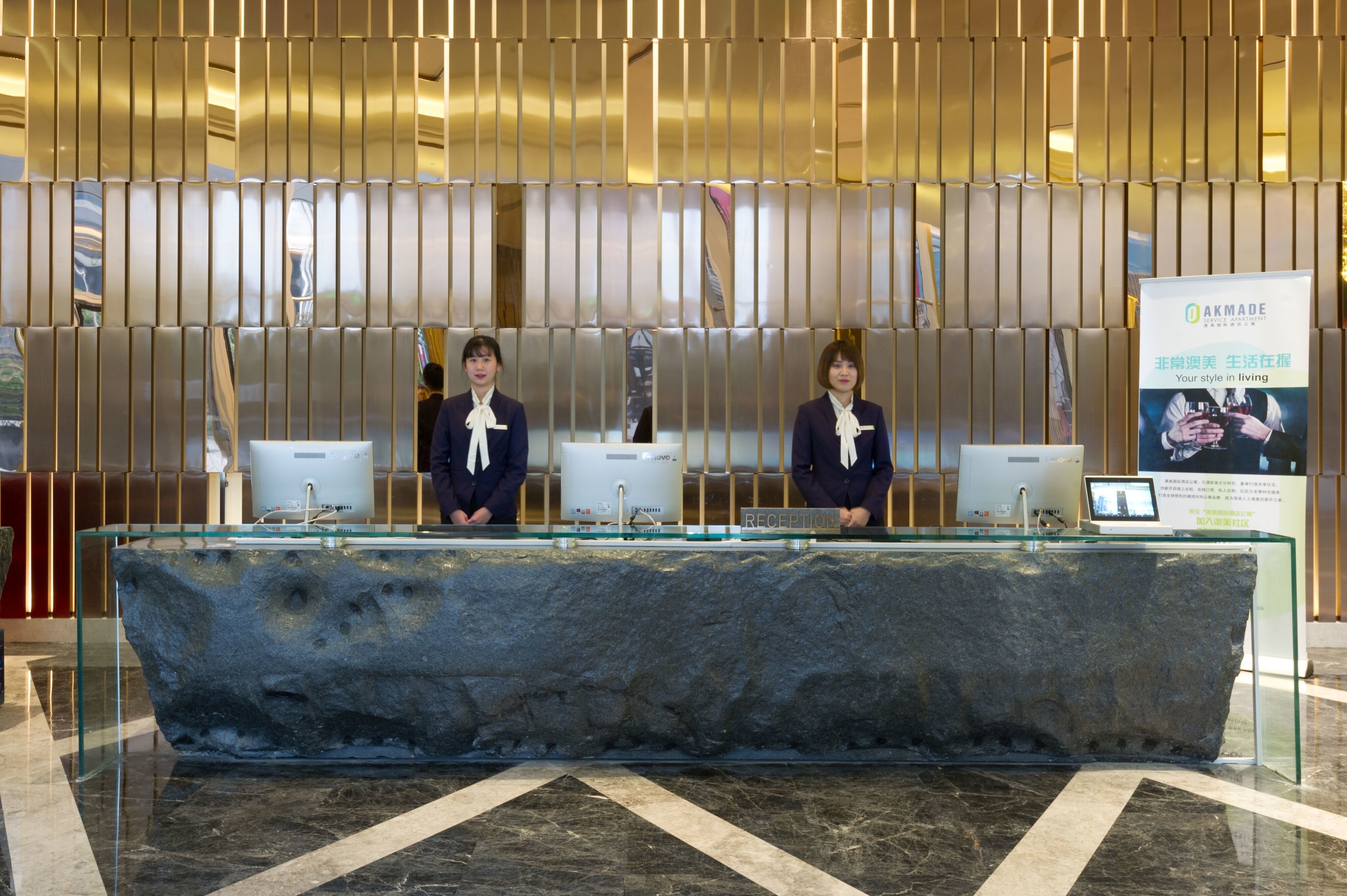 A reception desk with two staff members, a stone counter, and a modern interior with vertical blinds.