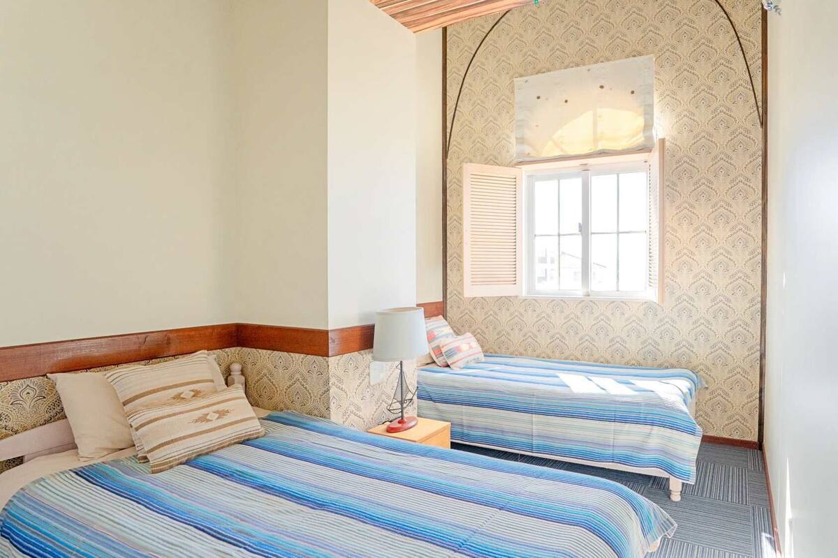 A bedroom with two beds, patterned wallpaper, a window with shutters, and a lamp on a bedside table.