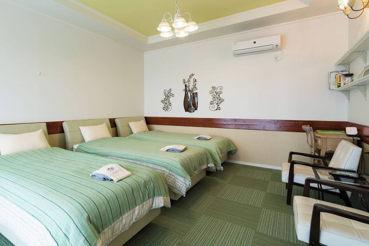 A hotel room with two beds, a desk, a chair, a shelf with books, and an air conditioning unit.