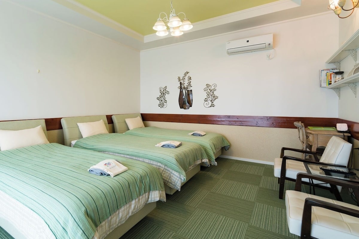 A hotel room with two beds, a desk, a chair, a shelf with books, and an air conditioning unit.
