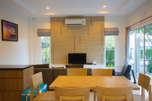 A modern dining area with a wooden table and chairs, a flat-screen TV, and a large window with a view of greenery.