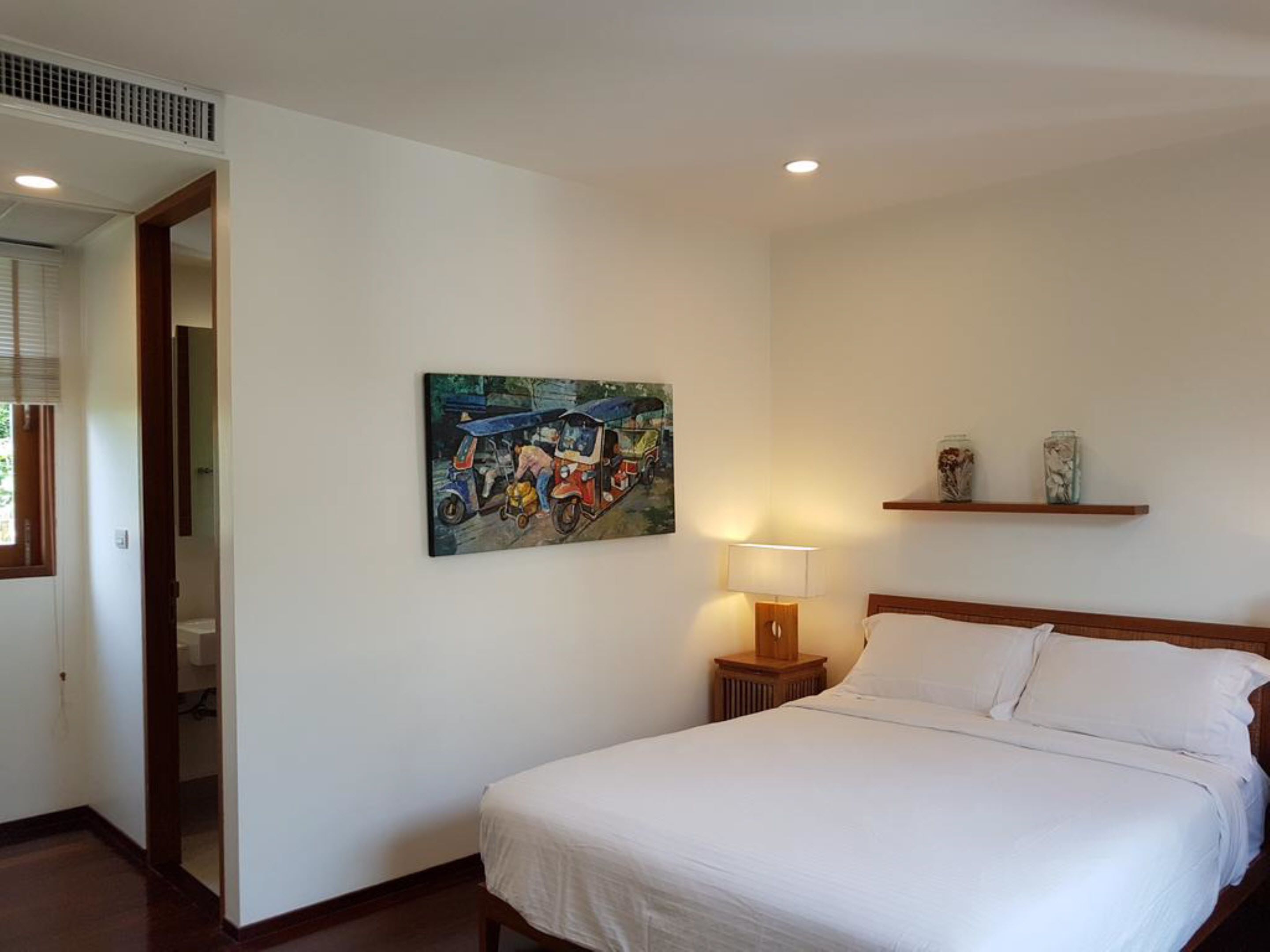 A bedroom with a bed, a nightstand with a lamp, a painting on the wall, and a shelf with decorative items.