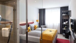 Habitació d'hotel amb dutxa envidriada, llit amb roba blanca i accents grocs, escriptori amb cadira i televisió sobre un suport.