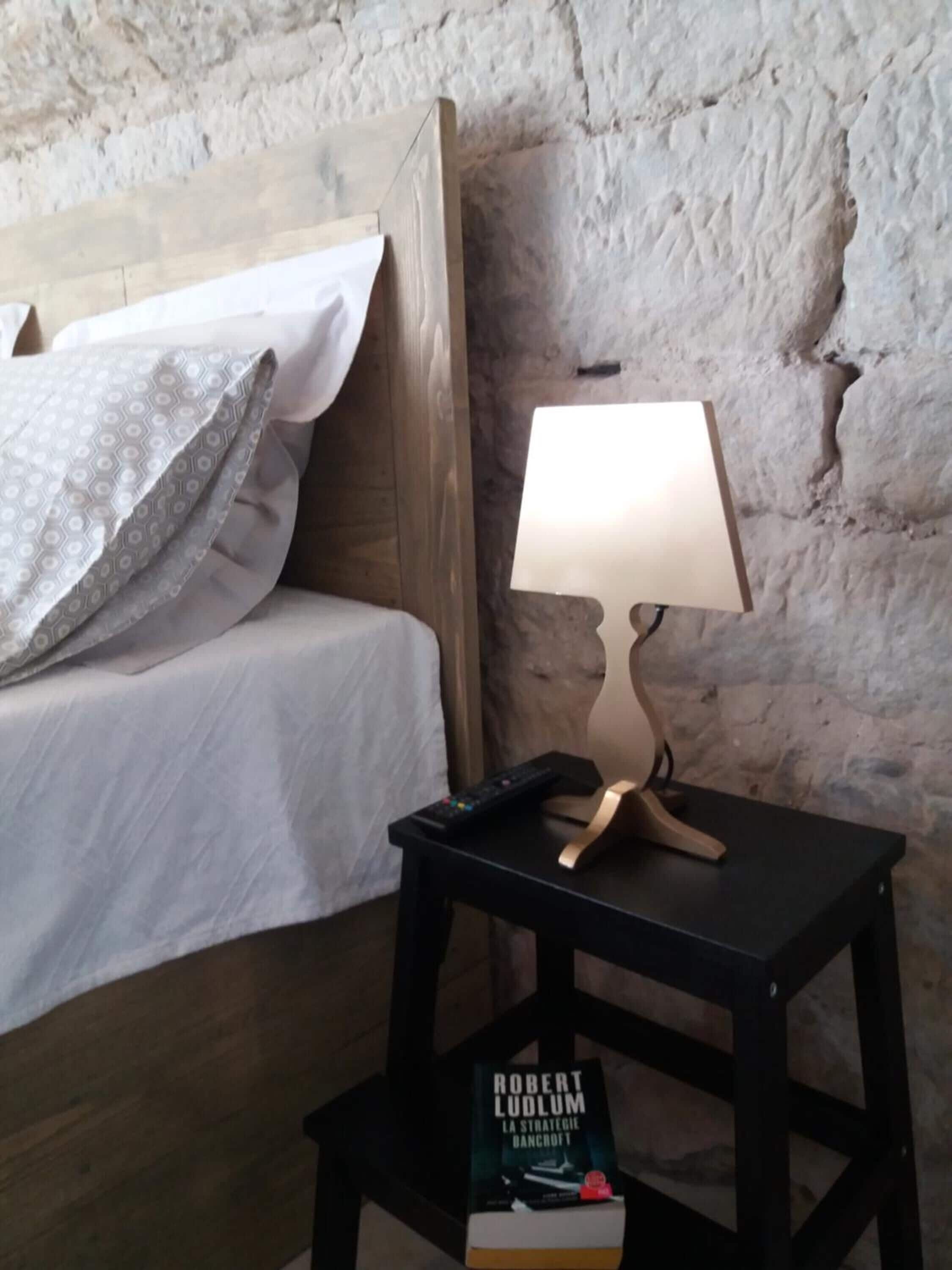 A bed with a wooden headboard, a bedside table with a lamp, and a book titled "La Strategie Bancroft" by Robert Ludlum.